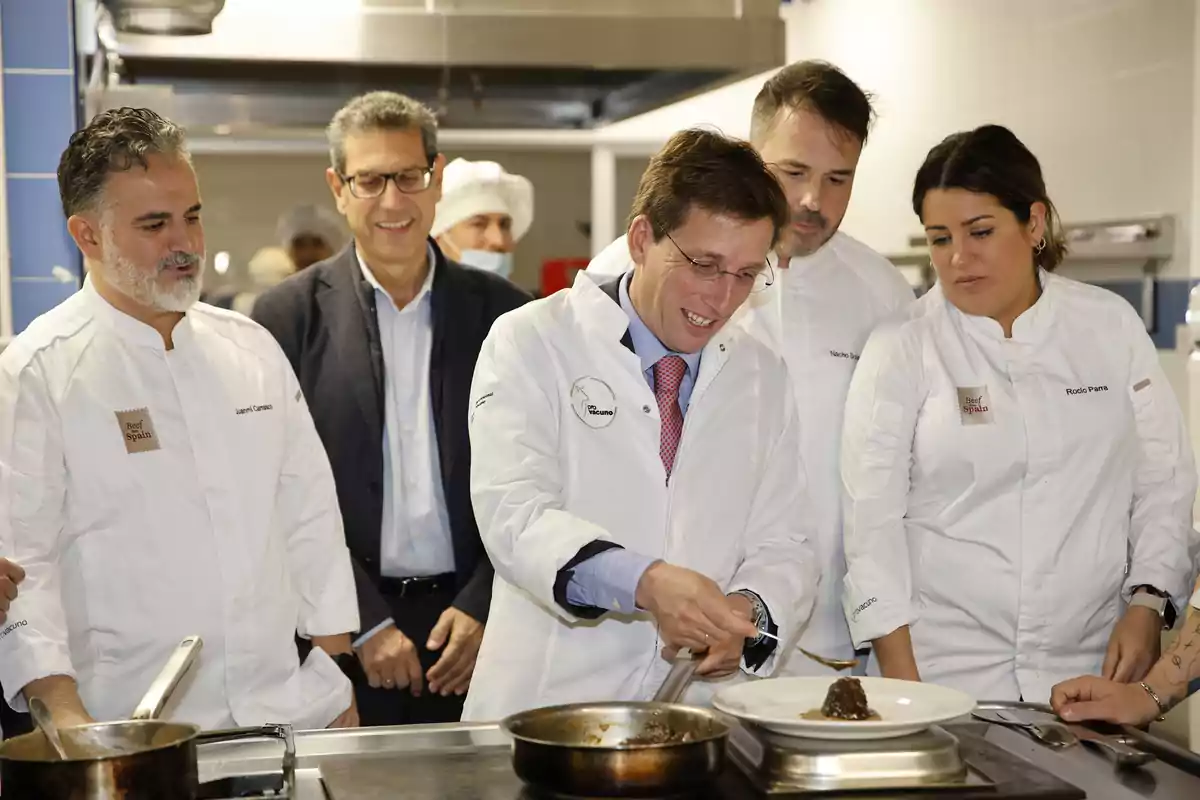 Un grupo de personas en una cocina profesional, algunas con chaquetas de chef, observan a un hombre sirviendo comida en un plato.