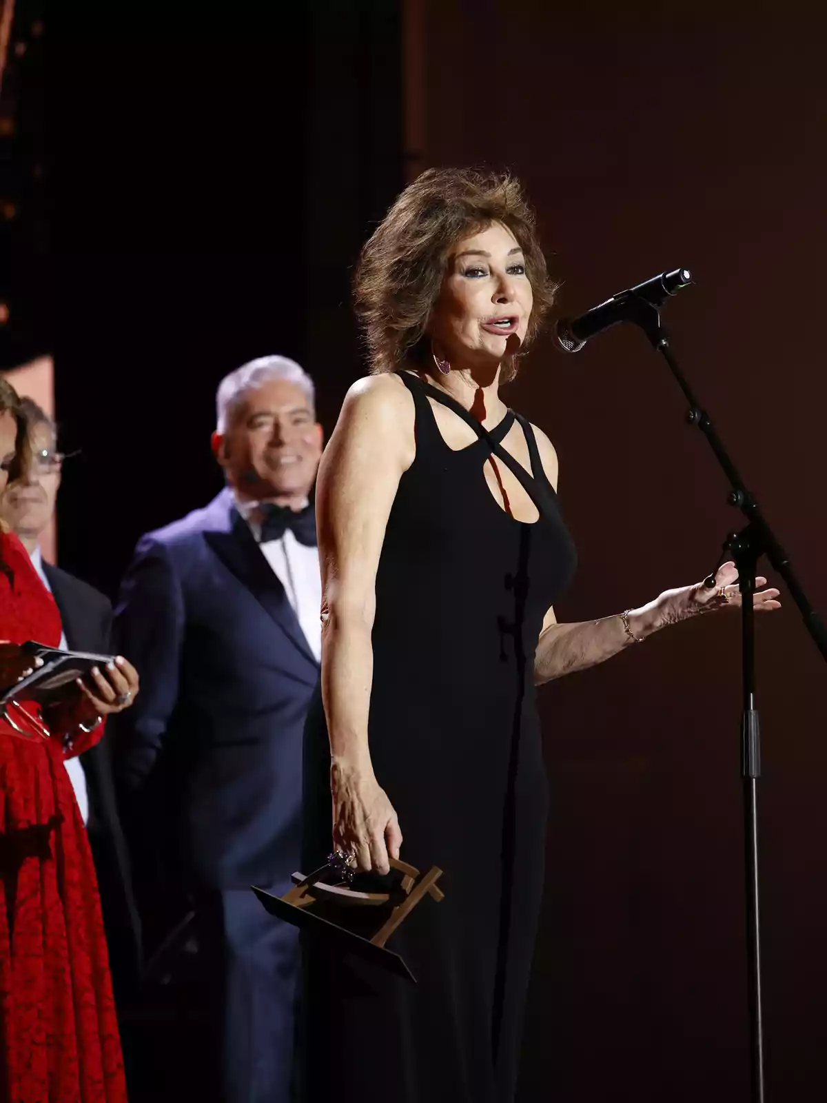 Una mujer con un vestido negro habla en un micrófono mientras sostiene un premio, con personas de fondo.