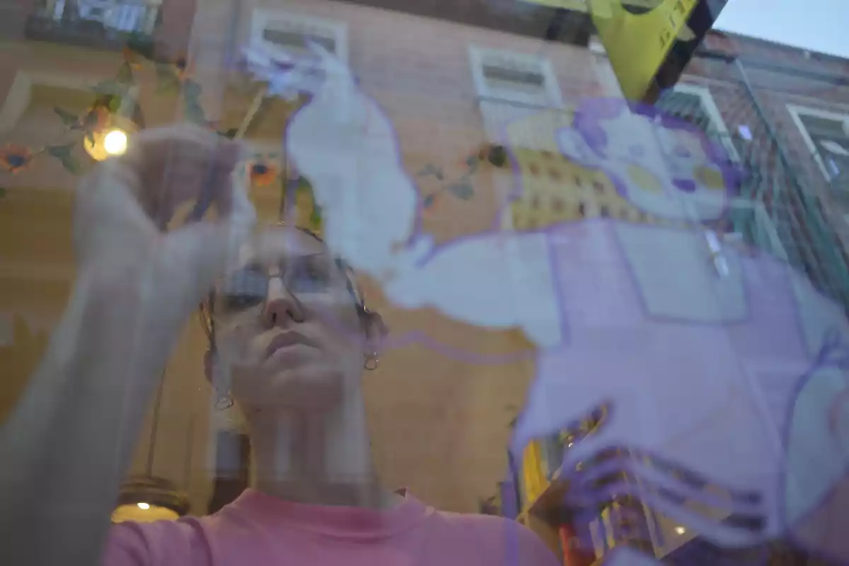 Una persona con gafas pinta un dibujo en una ventana desde el interior de un edificio reflejando el exterior y la decoración del lugar.