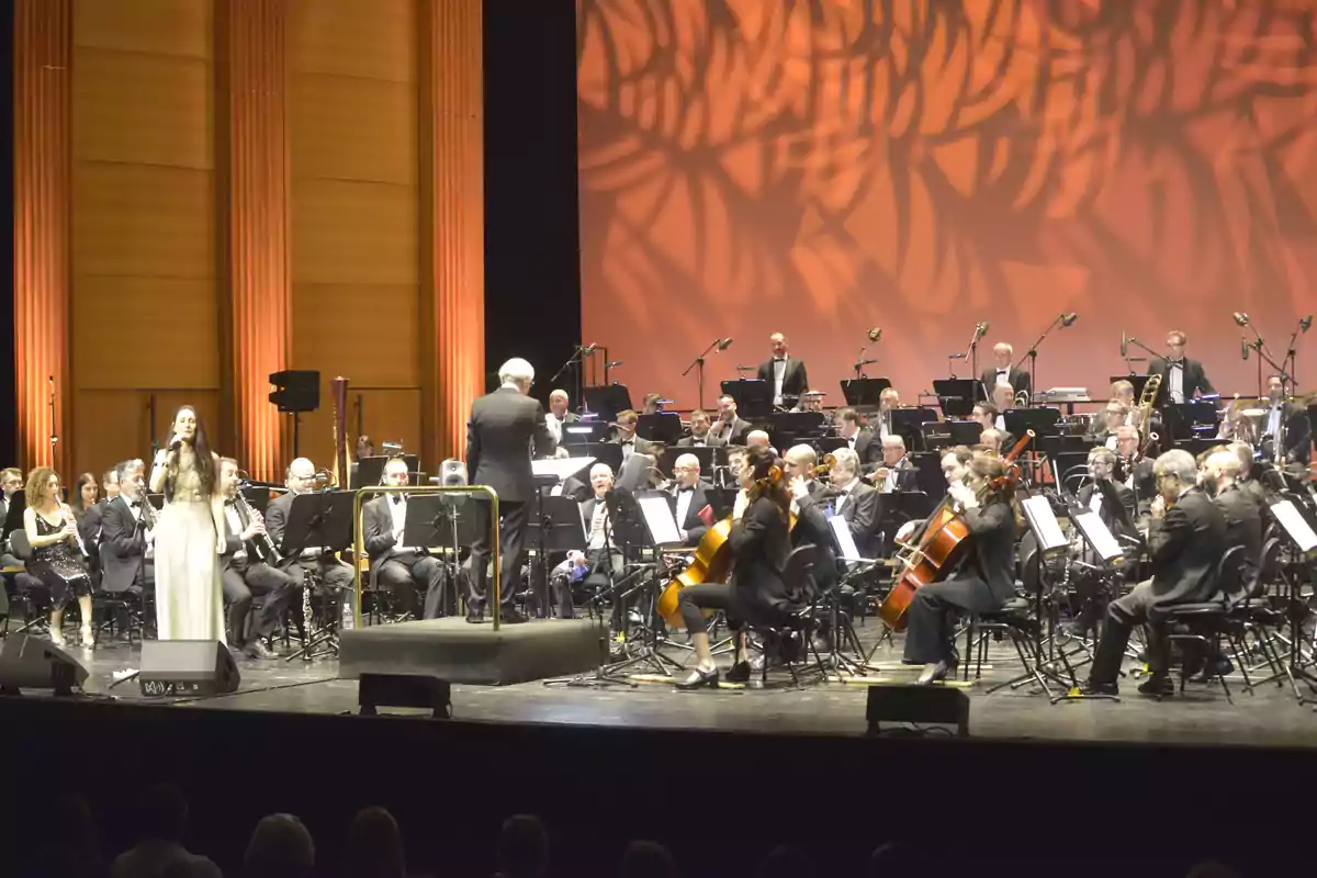 Una orquesta sinfónica en el escenario con una cantante solista y un director dirigiendo la música en un auditorio iluminado.