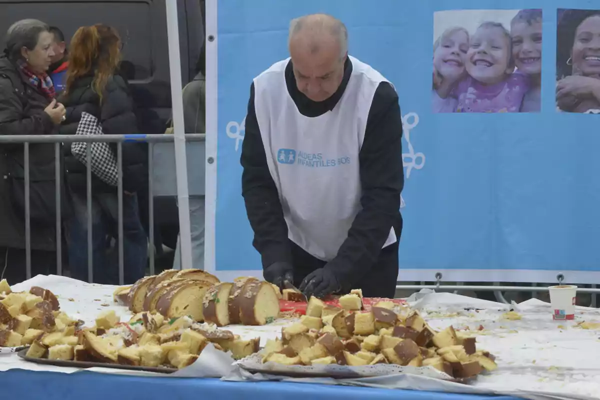 Un hombre corta un pastel grande en una mesa al aire libre mientras varias personas observan, con un cartel de Aldeas Infantiles SOS de fondo.