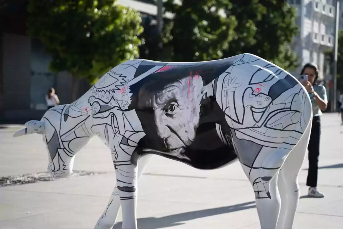 Escultura de una vaca decorada con un retrato en blanco y negro de un hombre y dibujos abstractos en un espacio público al aire libre.