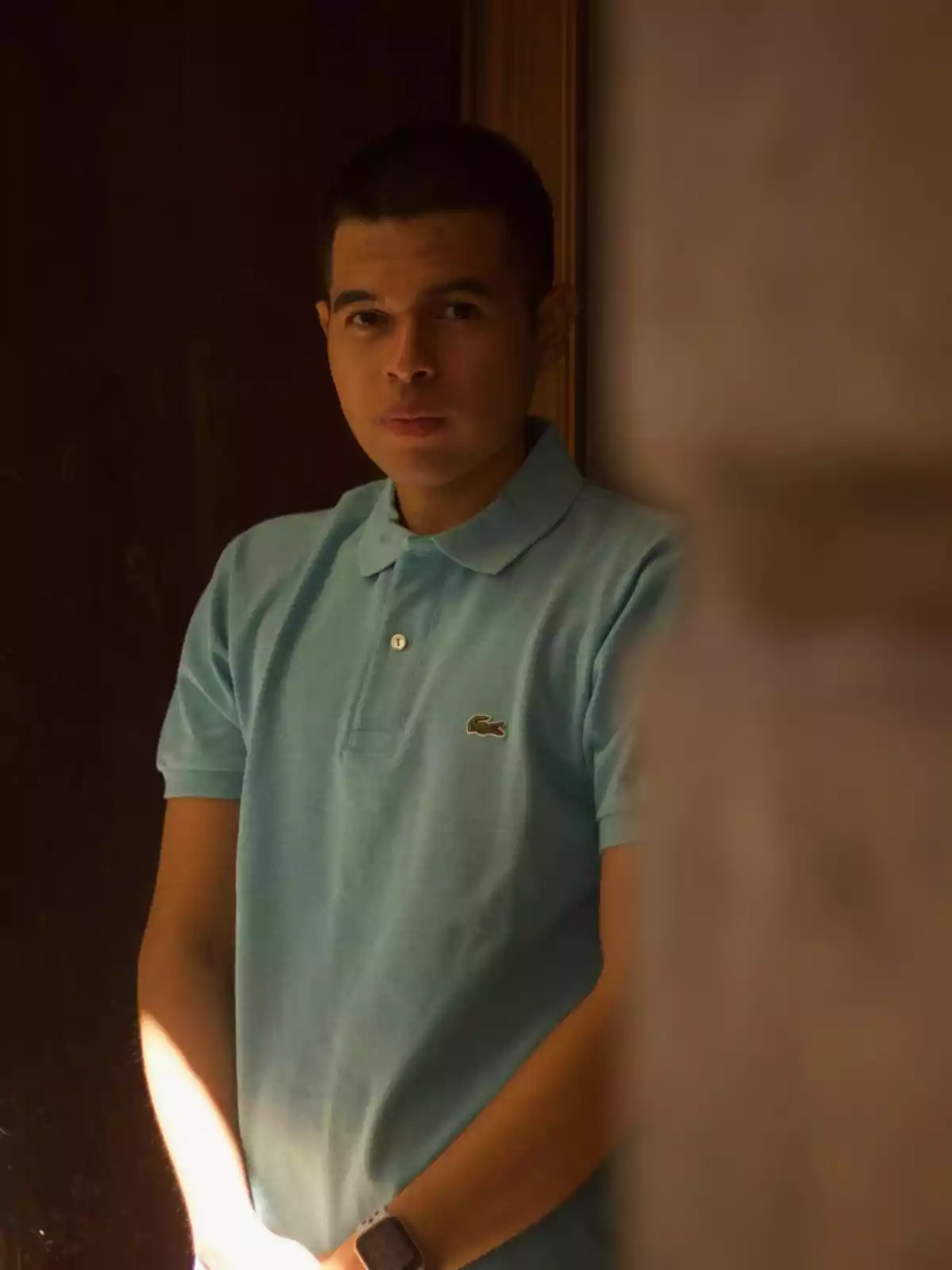 Un hombre con una camiseta polo azul claro y un reloj de pulsera, de pie en un entorno oscuro con una expresión seria.