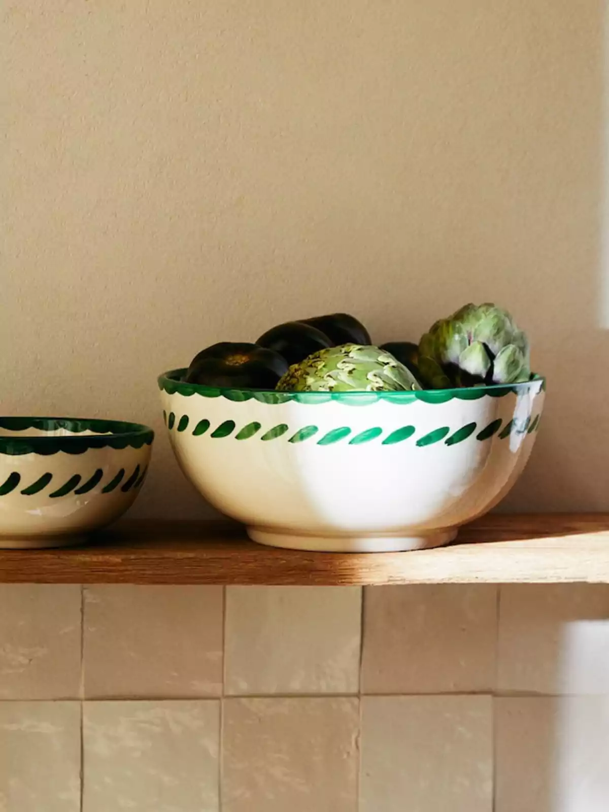 Un tazón de cerámica blanca con un borde decorativo verde, lleno de alcachofas y berenjenas, colocado sobre un estante de madera junto a otro tazón más pequeño con un diseño similar.