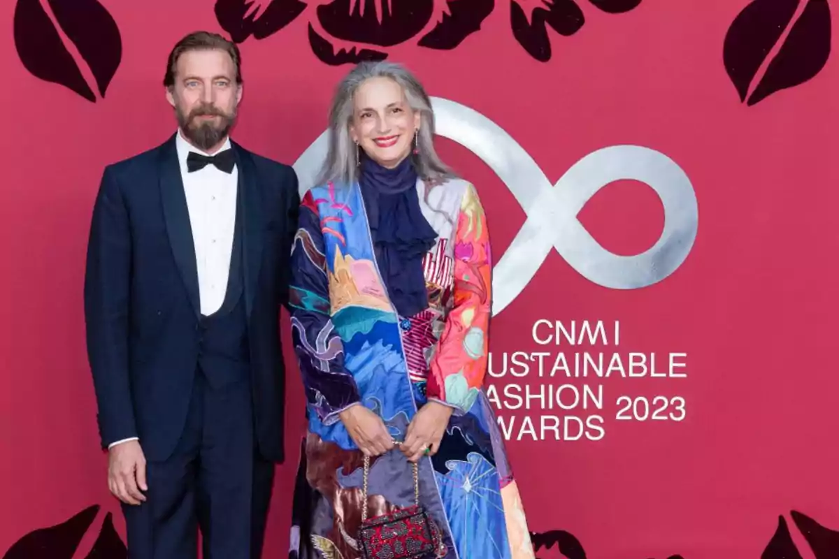 Un hombre con esmoquin oscuro y una mujer con un abrigo colorido posan juntos frente a un fondo rojo con el logo y texto de los CNMI Sustainable Fashion Awards 2023. Un hombre con esmoquin oscuro y una mujer con un abrigo colorido posan juntos frente a un fondo rojo con el logo y texto de los CNMI Sustainable Fashion Awards 2023.