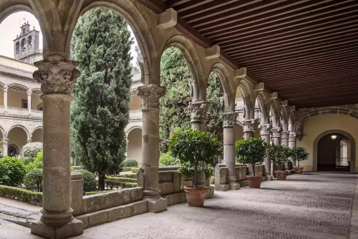 Claustro de un edificio histórico con columnas de piedra y arcos, rodeado de vegetación y árboles, con un pasillo techado y un jardín central.