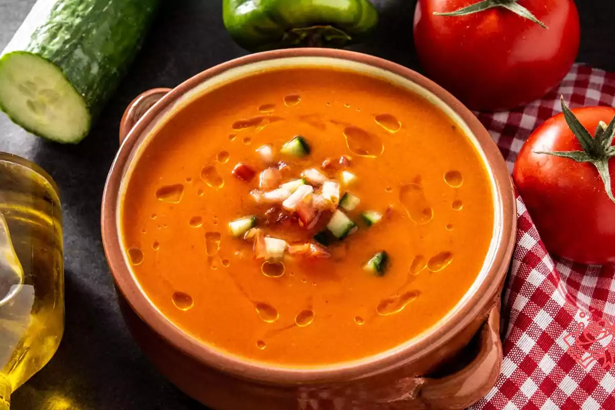 Un plato de gazpacho con trozos de pepino, tomate y cebolla, rodeado de ingredientes frescos como pepino, pimiento verde y tomates, sobre un mantel a cuadros rojos y blancos.