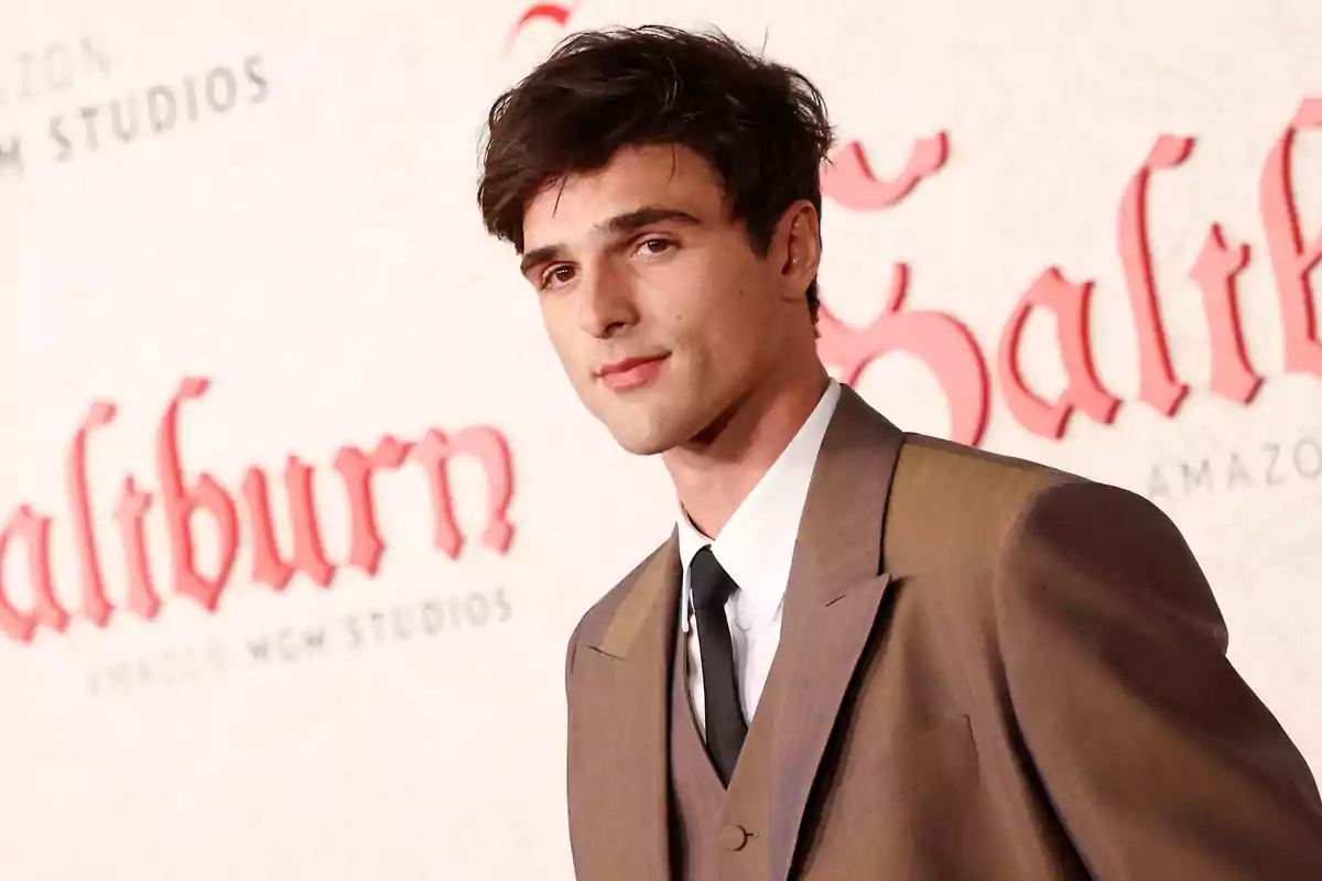 Hombre joven con cabello oscuro y traje marrón posando en una alfombra roja frente a un fondo con letras rojas