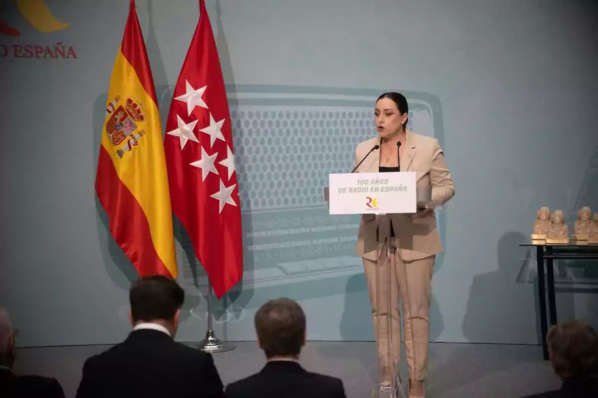 Una persona hablando en un podio durante un evento conmemorativo de los 100 años de la radio en España, con las banderas de España y de la Comunidad de Madrid al fondo.