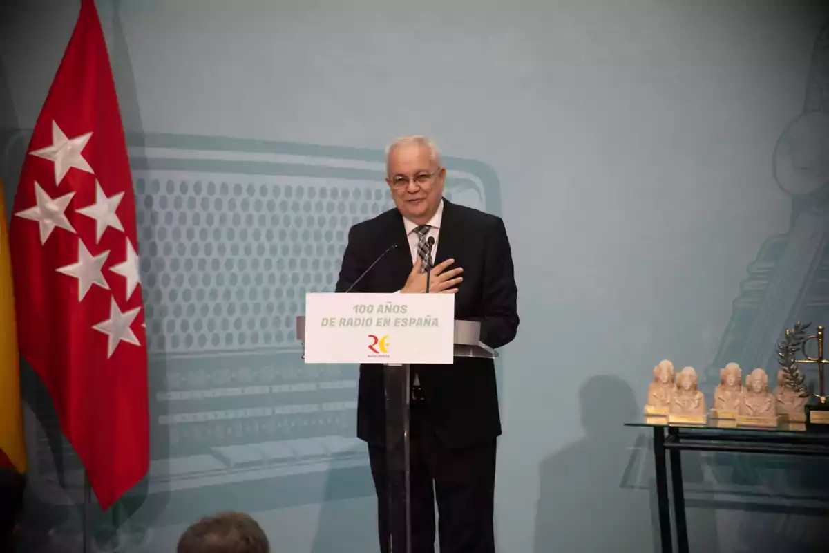 Un hombre hablando en un podio durante un evento de celebración de los 100 años de la radio en España, con una bandera roja a su lado y varios premios sobre una mesa.