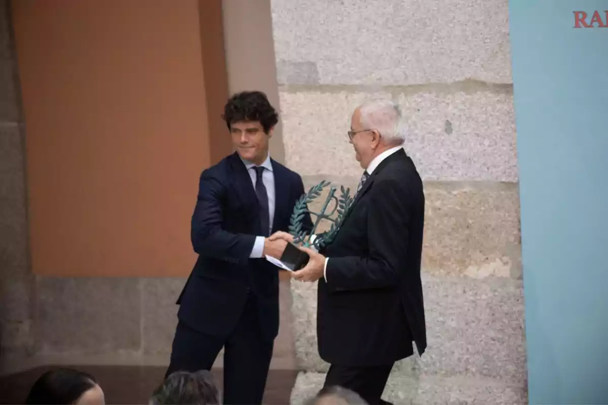 Dos hombres de traje se dan la mano durante una ceremonia de entrega de premios en un entorno formal.