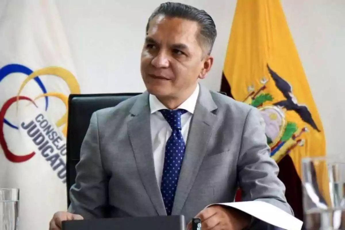 Un hombre con traje gris y corbata azul está sentado en una oficina con una bandera de Ecuador y un cartel del Consejo de la Judicatura en el fondo.