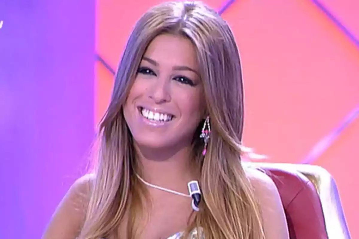 Una mujer sonriente con cabello largo y rubio sentada en un estudio de televisión con fondo rosa. Una mujer sonriente con cabello largo y rubio sentada en un estudio de televisión con fondo rosa.