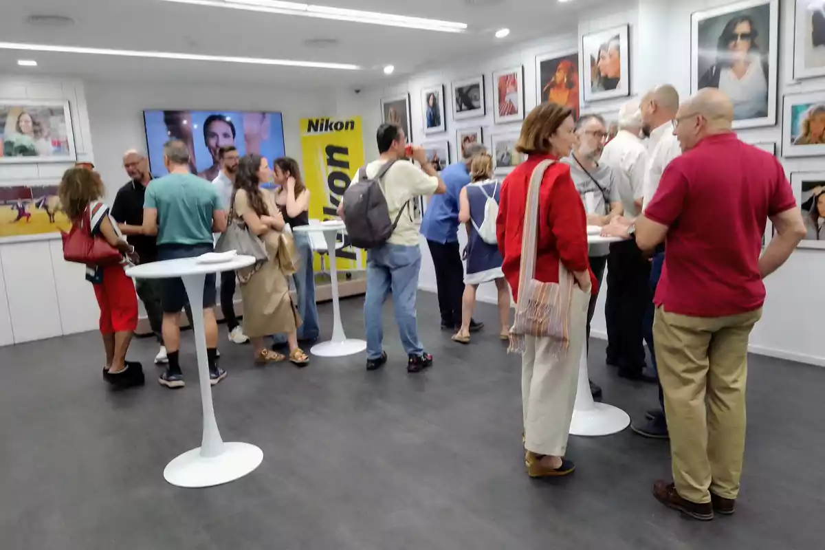 Personas conversando y observando fotografías en una galería con mesas altas y un ambiente moderno Personas conversando y observando fotografías en una galería con mesas altas y un ambiente moderno
