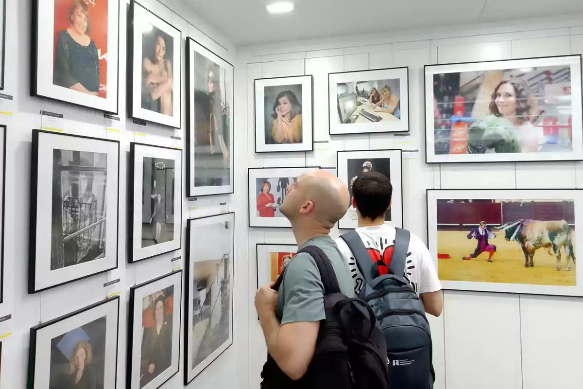 Dos personas con mochilas observan atentamente una exposición de fotografías enmarcadas en una galería de arte