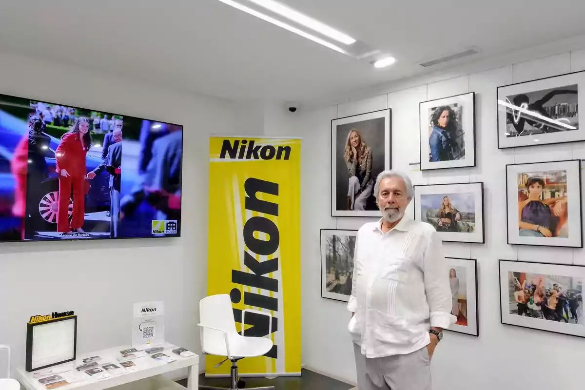 Hombre de cabello canoso y barba de pie en una galería de fotos con un banner amarillo de Nikon y una pantalla mostrando una imagen de una mujer vestida de rojo Hombre de cabello canoso y barba de pie en una galería de fotos con un banner amarillo de Nikon y una pantalla mostrando una imagen de una mujer vestida de rojo
