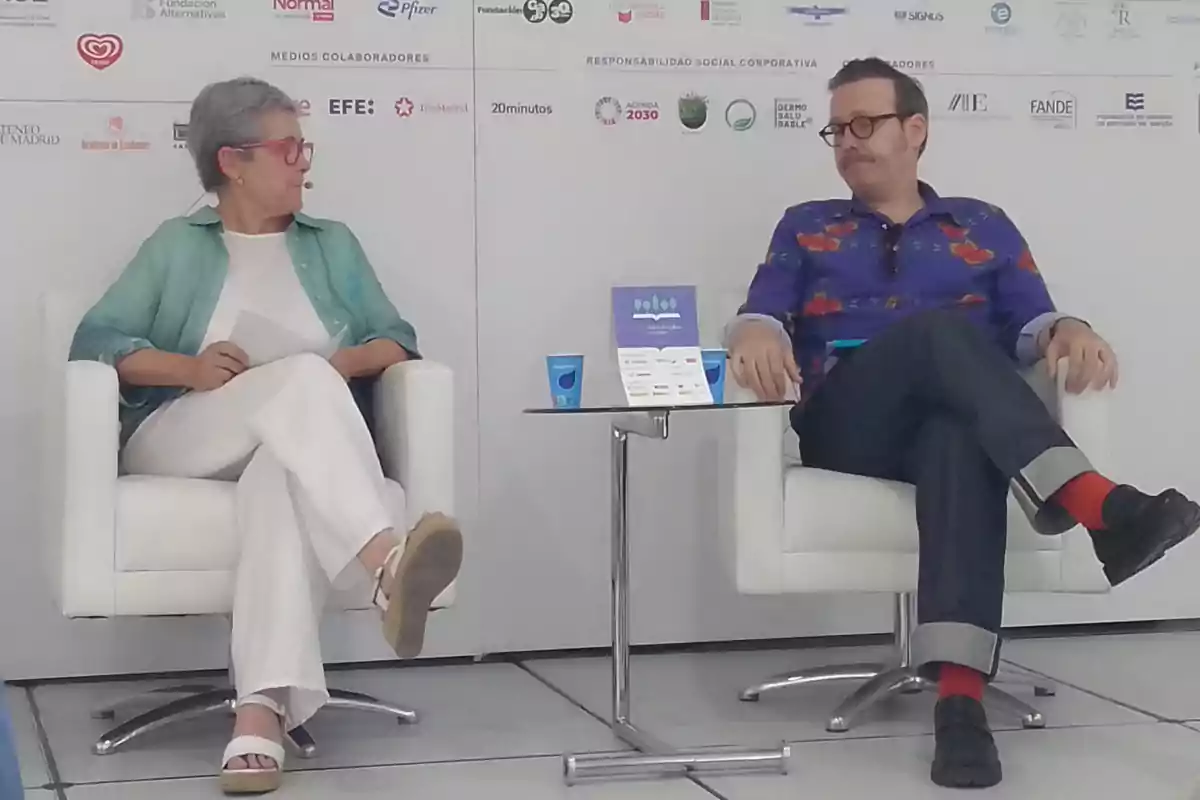 Dos personas sentadas en sillones blancos conversan en un escenario con fondo de logotipos de patrocinadores y una mesa pequeña entre ellas Dos personas sentadas en sillones blancos conversan en un escenario con fondo de logotipos de patrocinadores y una mesa pequeña entre ellas