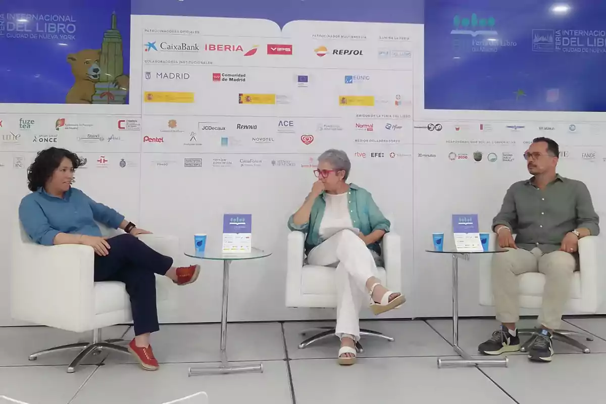Tres personas sentadas en sillones blancos participan en una charla en un evento literario, con un fondo lleno de logotipos de patrocinadores y colaboradores.