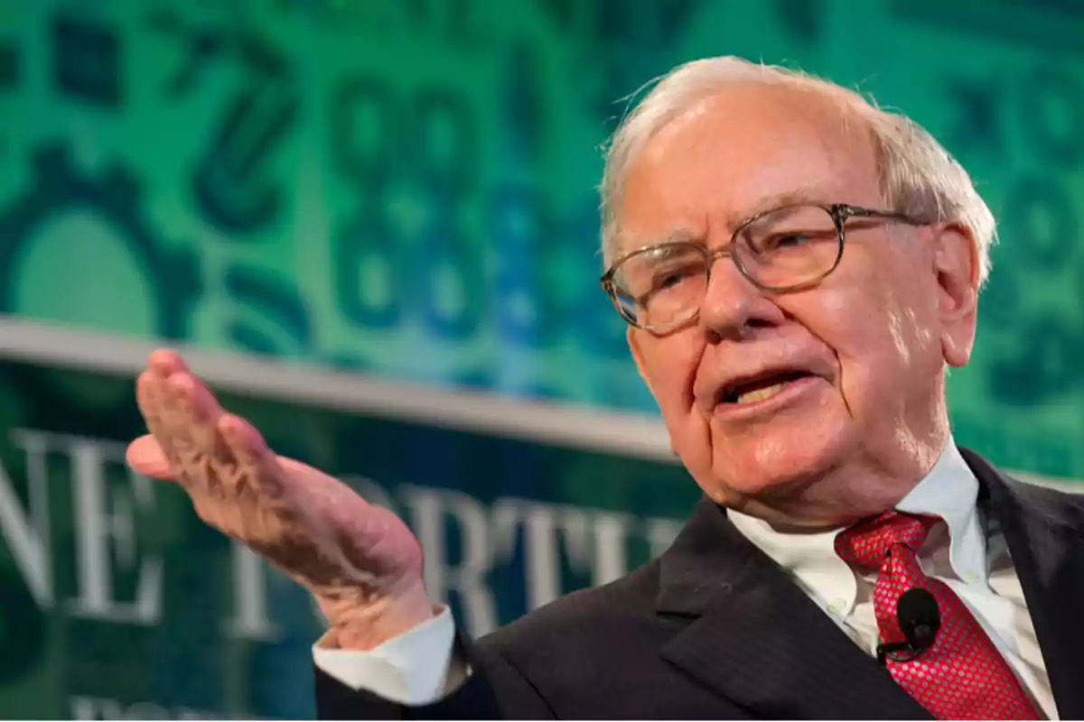 An older man with glasses and a red tie gestures while speaking at an event.