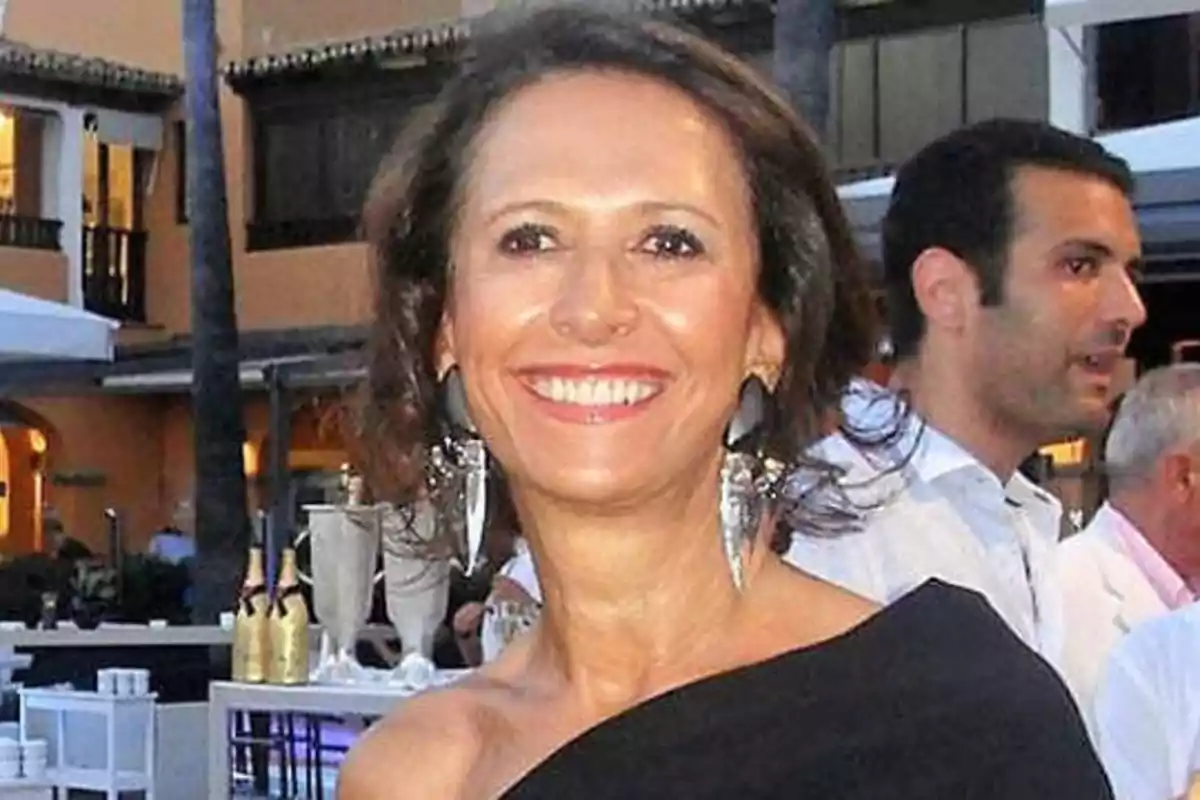 Una mujer sonriente con un vestido negro y pendientes grandes en un evento al aire libre.