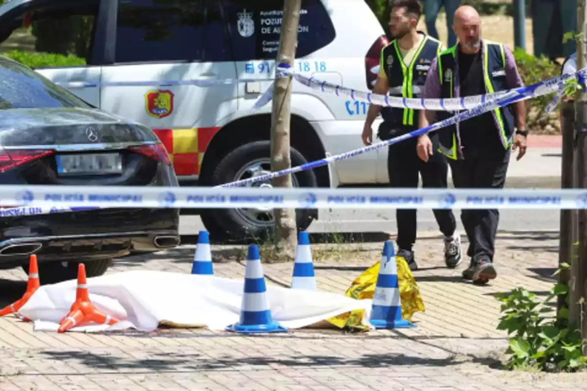 Dos agentes de policía caminan cerca de un área acordonada con cinta policial donde hay un cuerpo cubierto por una sábana blanca en la acera junto a un coche negro y varios conos de tráfico.