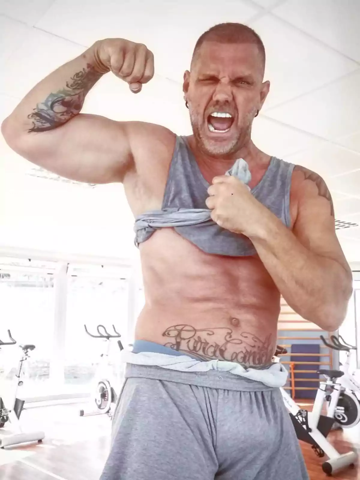 Un hombre musculoso con tatuajes en el brazo y el abdomen, levantando su camiseta y flexionando el bíceps en un gimnasio con bicicletas estáticas al fondo.