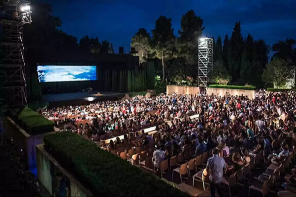 Una multitud de personas sentadas al aire libre viendo una proyección en una pantalla grande durante la noche.