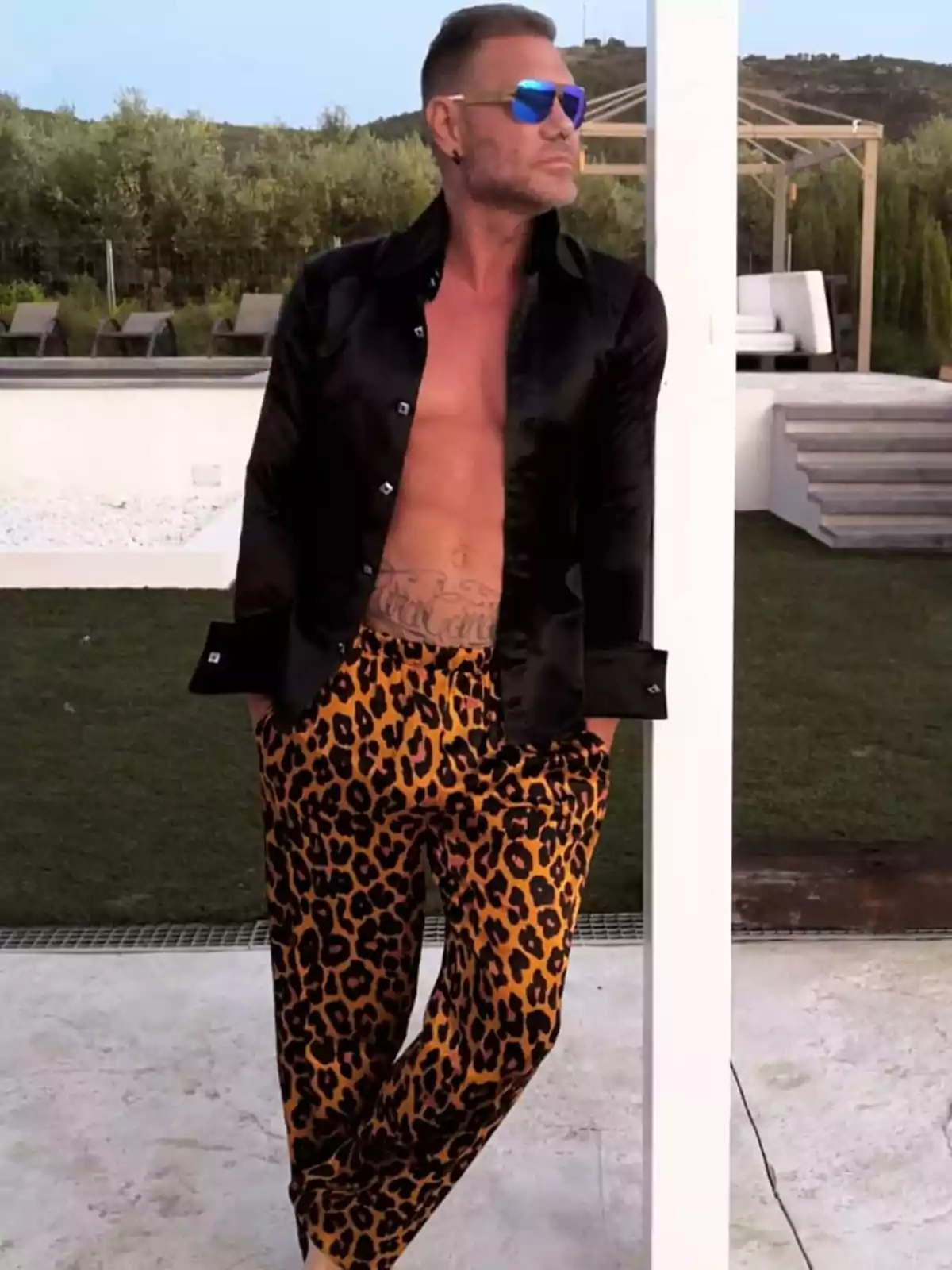 Hombre con camisa negra abierta y pantalones de estampado de leopardo, apoyado en una columna al aire libre.