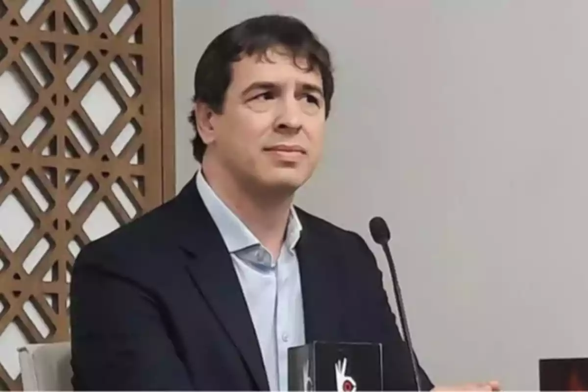 Un hombre con traje oscuro y camisa clara está sentado frente a un micrófono en un entorno interior con un panel decorativo de fondo.