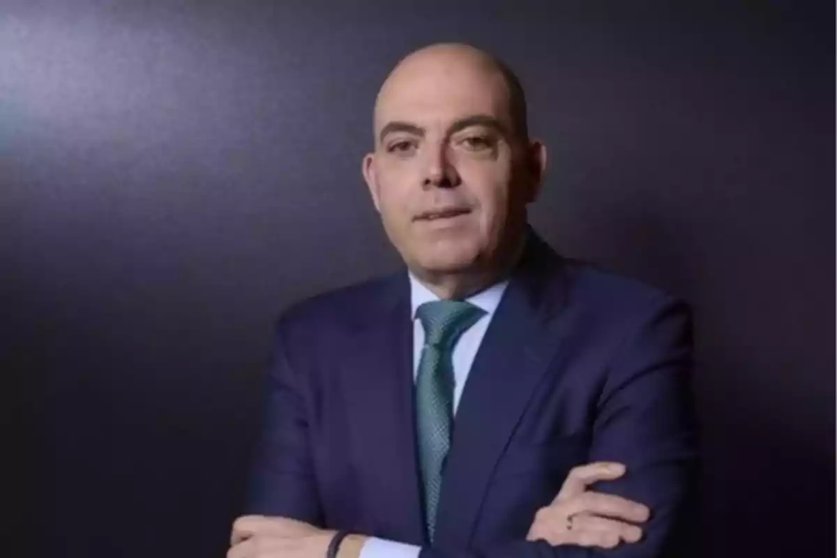 Un hombre calvo con traje azul y corbata verde posa con los brazos cruzados frente a un fondo oscuro. Un hombre calvo con traje azul y corbata verde posa con los brazos cruzados frente a un fondo oscuro.