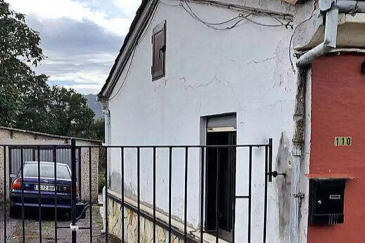 Una casa blanca con una puerta y una ventana pequeñas, un buzón negro en la pared y un coche azul estacionado detrás de una reja.