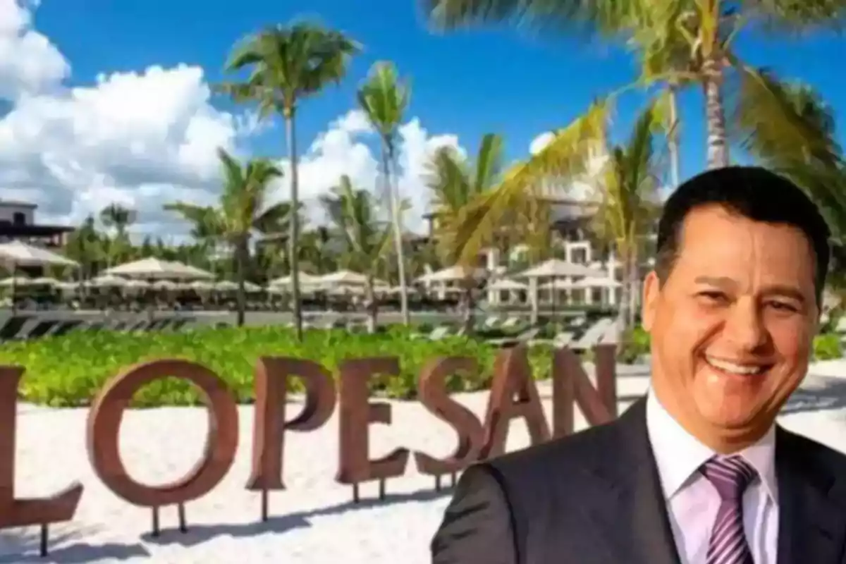 Hombre sonriente con traje frente a un letrero grande de madera que dice Lopesan en una playa con palmeras y cielo azul Hombre sonriente con traje frente a un letrero grande de madera que dice Lopesan en una playa con palmeras y cielo azul