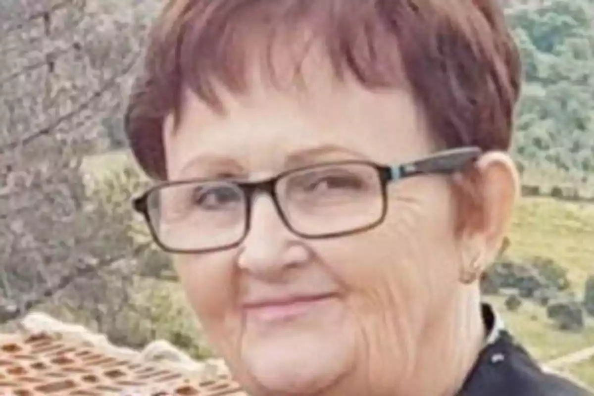 Una mujer mayor con gafas y cabello corto castaño claro sonríe mientras está al aire libre con un paisaje natural de fondo.