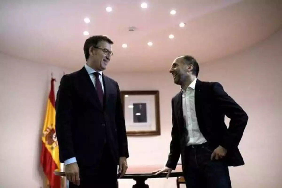 Dos hombres de traje conversan y ríen en una sala con una bandera de España en el fondo.