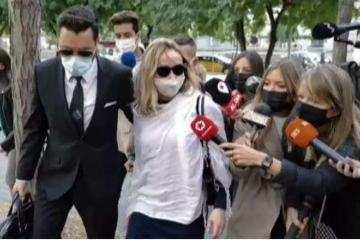 Una mujer con gafas de sol y mascarilla es escoltada por un hombre con traje mientras varios periodistas con micrófonos la rodean.