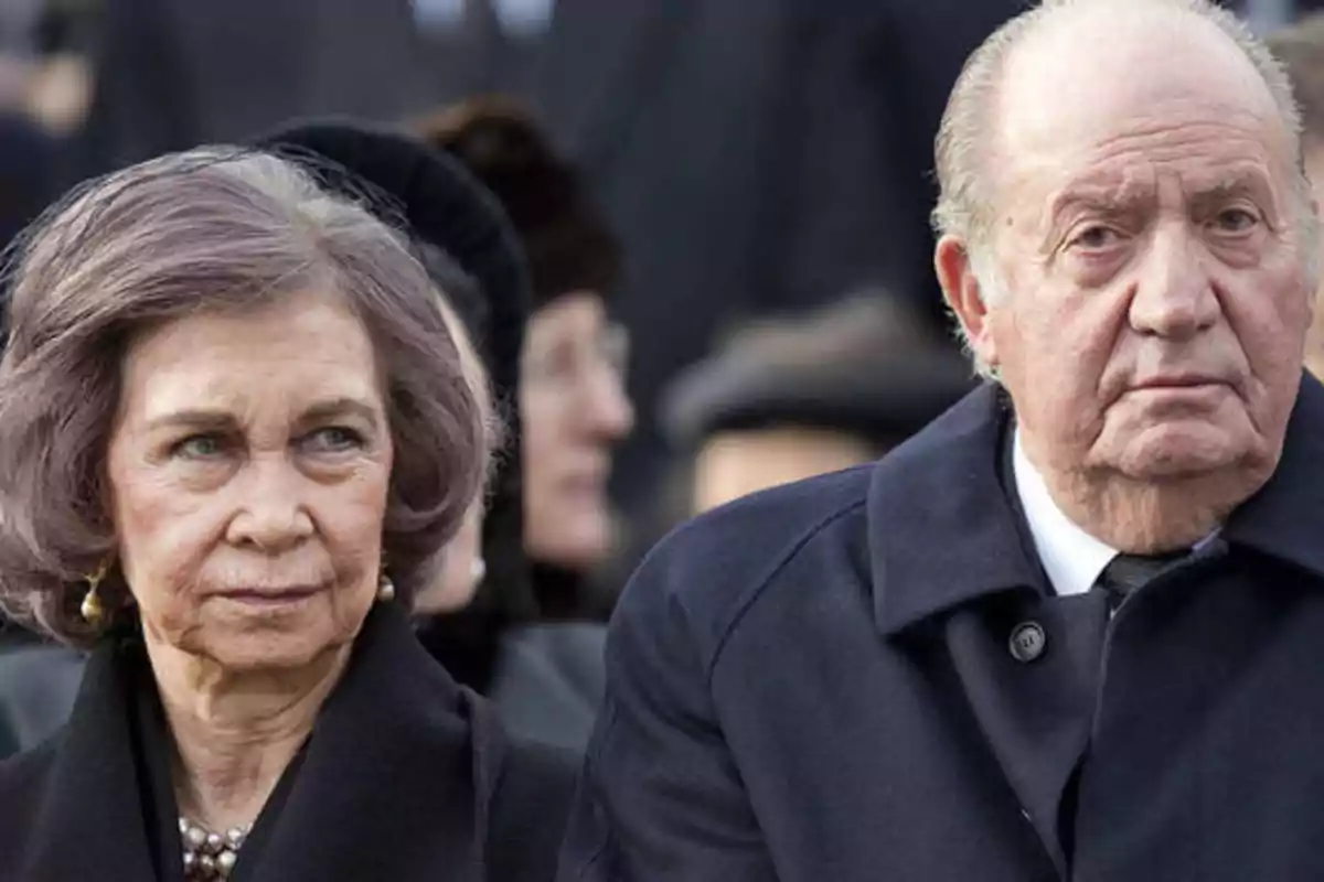 Dos personas mayores con expresión seria, vestidas con abrigos oscuros, en un evento formal al aire libre.