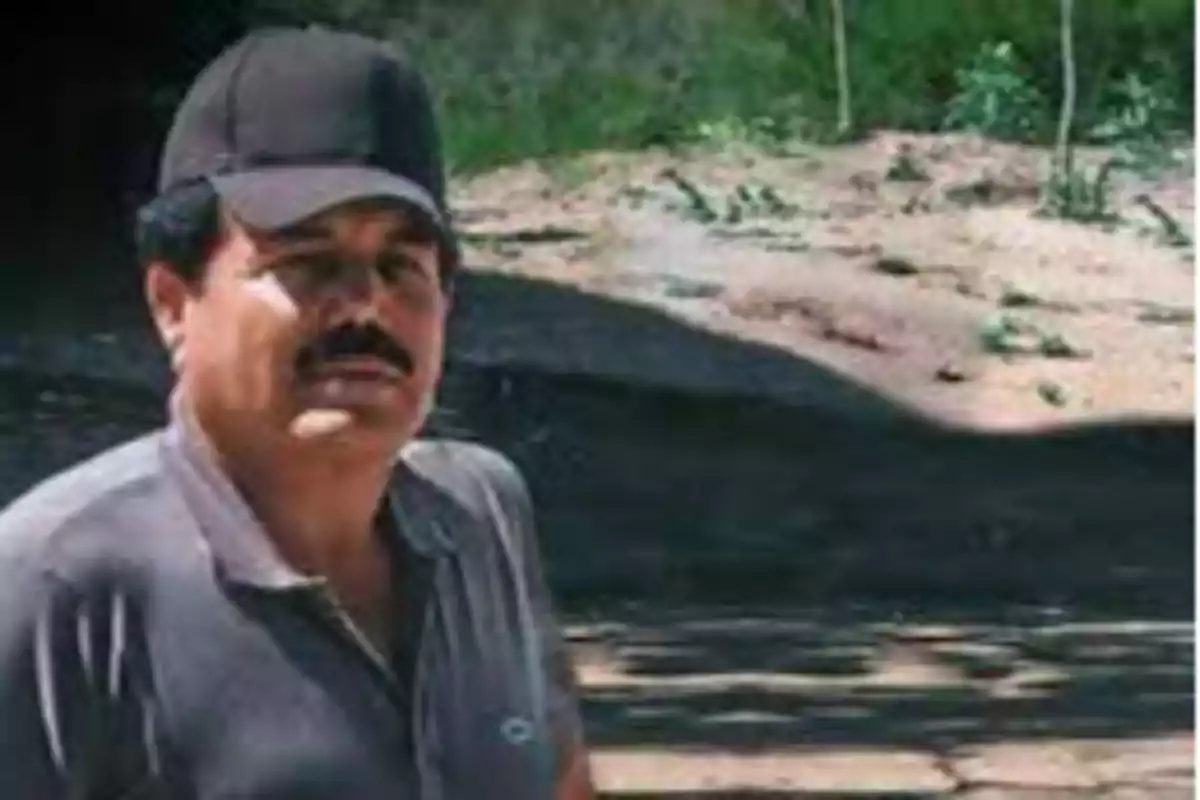 Un hombre con bigote y gorra negra está de pie al aire libre en un entorno natural.