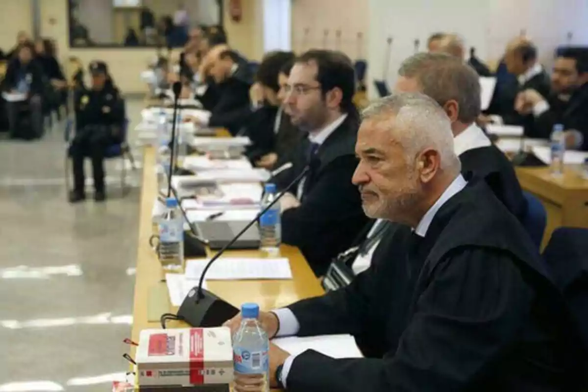 Un grupo de personas, algunas con toga, están sentadas en una mesa larga con documentos y botellas de agua, en lo que parece ser una sala de audiencias o un tribunal.