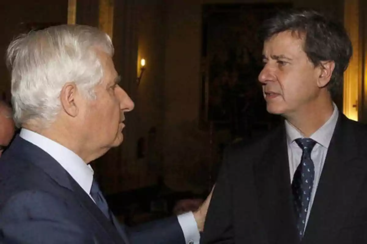 Dos hombres de traje conversando en un ambiente interior con poca luz.