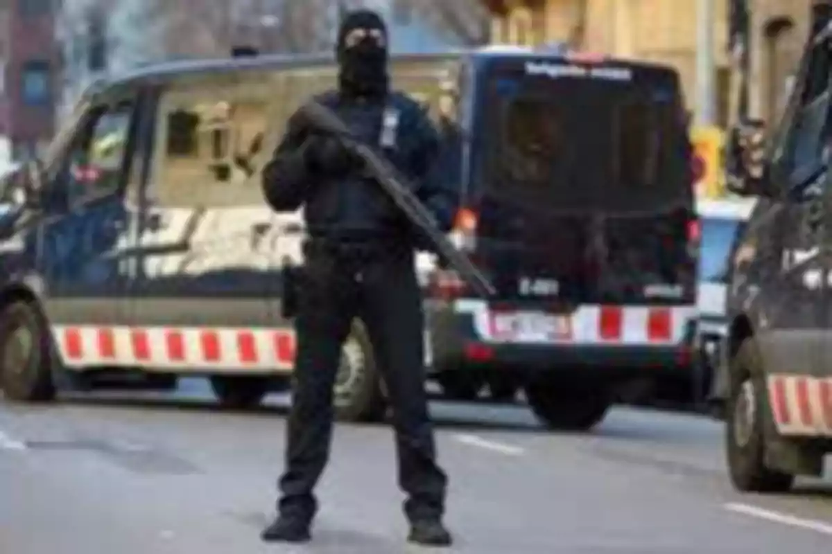 Un agente de policía con pasamontañas sostiene un arma frente a vehículos policiales en una calle.