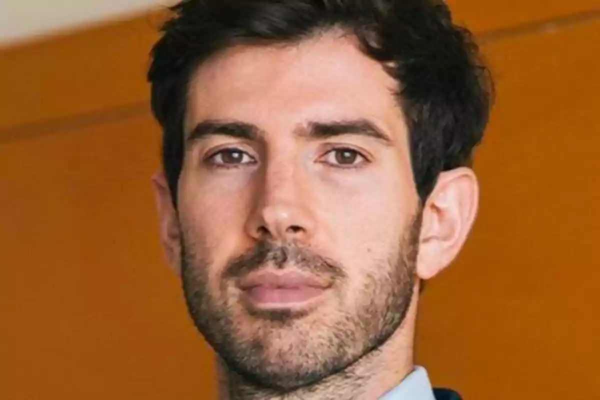 Hombre con barba y cabello oscuro frente a un fondo naranja.