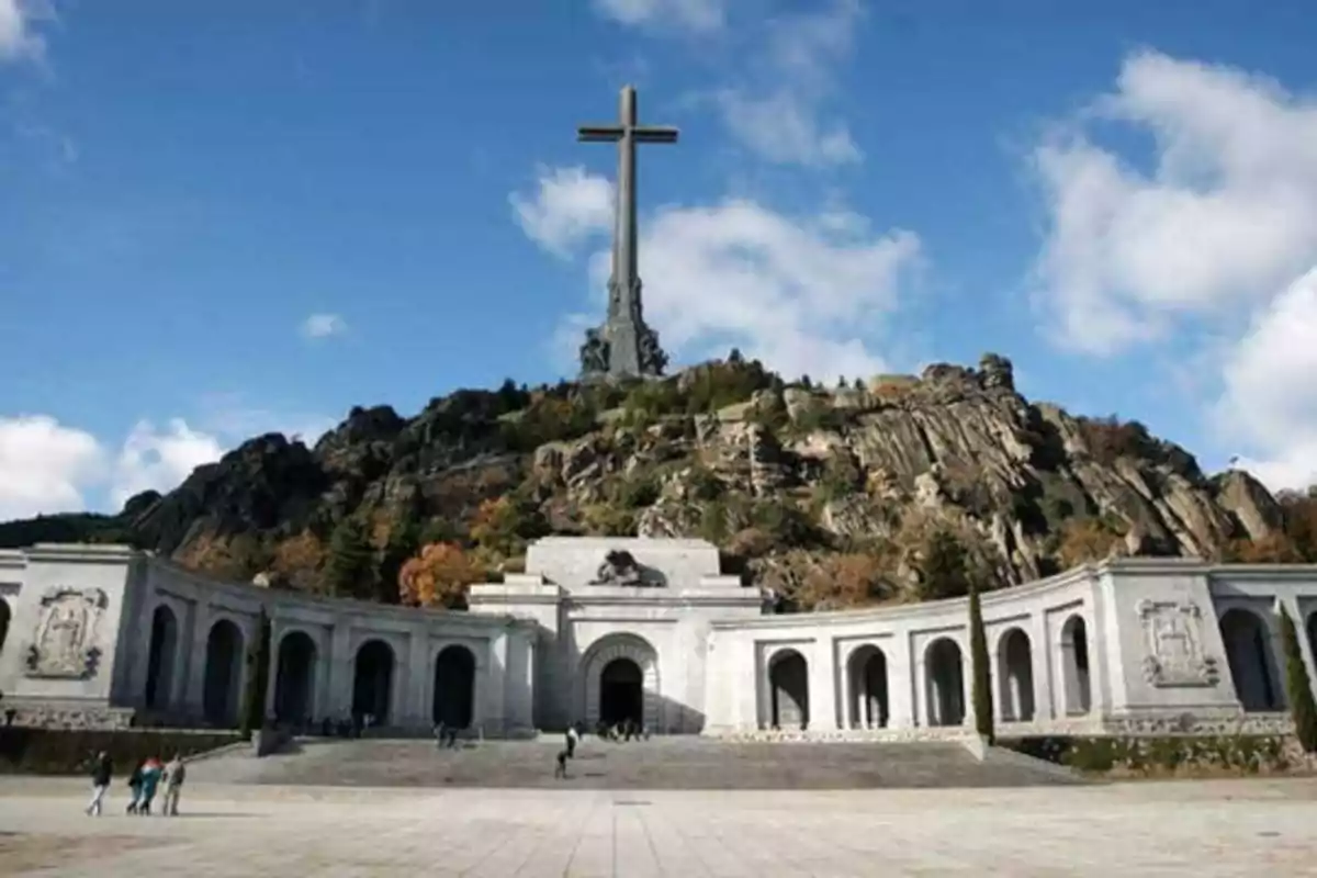 Una gran cruz de piedra se alza sobre una colina rocosa, con un edificio de arquitectura clásica en la base y un cielo azul con algunas nubes en el fondo.