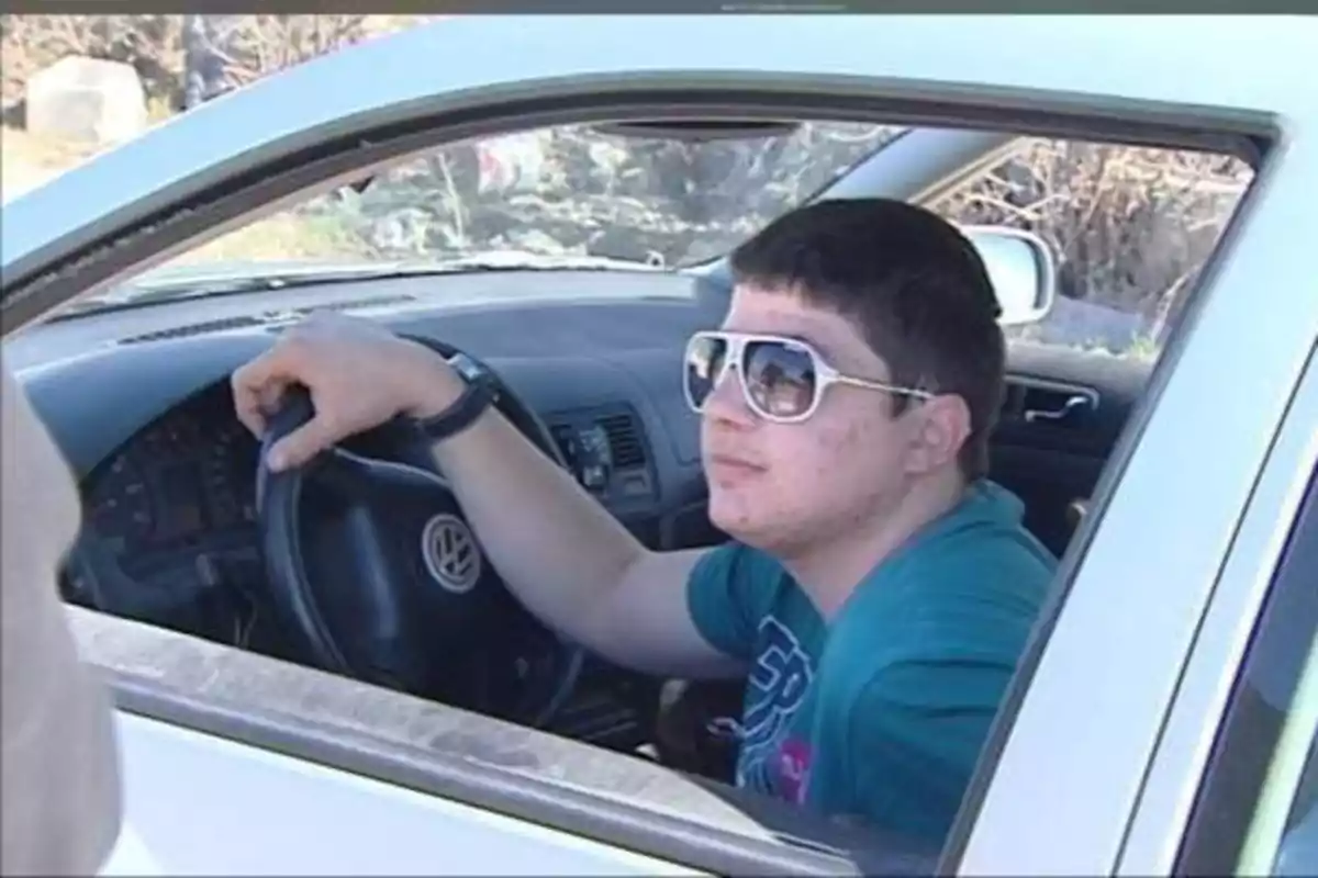 Un joven con gafas de sol sentado al volante de un coche blanco.