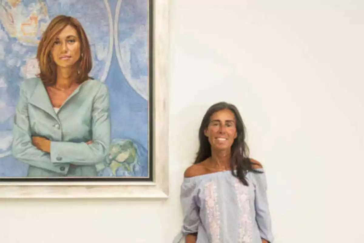 Una mujer de pie junto a un retrato de otra mujer en una pared blanca.