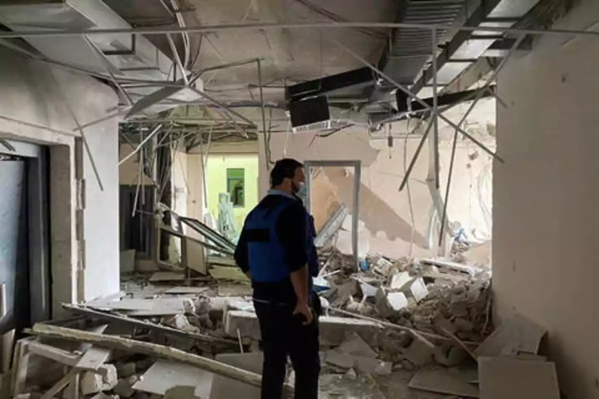 Una persona con chaleco azul y mascarilla observa los escombros y daños en el interior de un edificio.