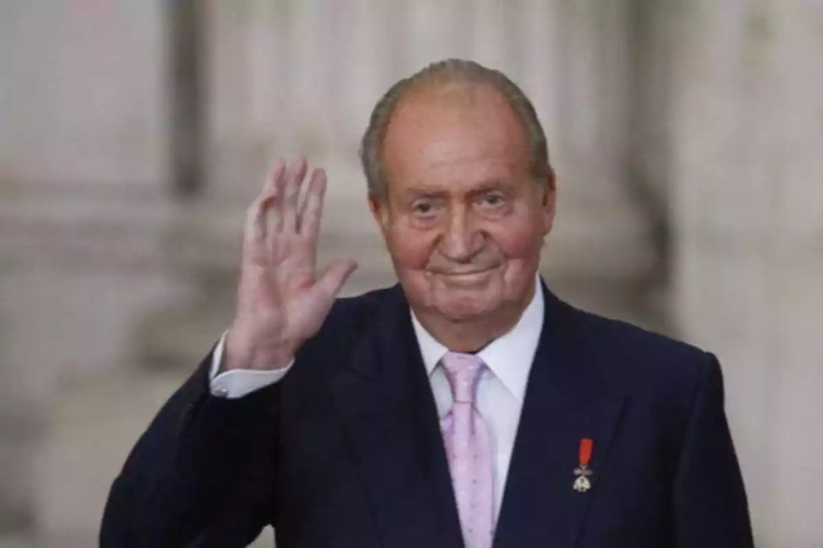 Hombre mayor con traje oscuro y corbata rosa saludando con la mano derecha levantada.