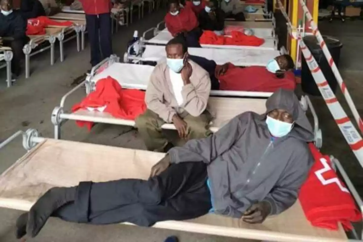 Varias personas con mascarillas descansan en camas improvisadas en un refugio temporal.