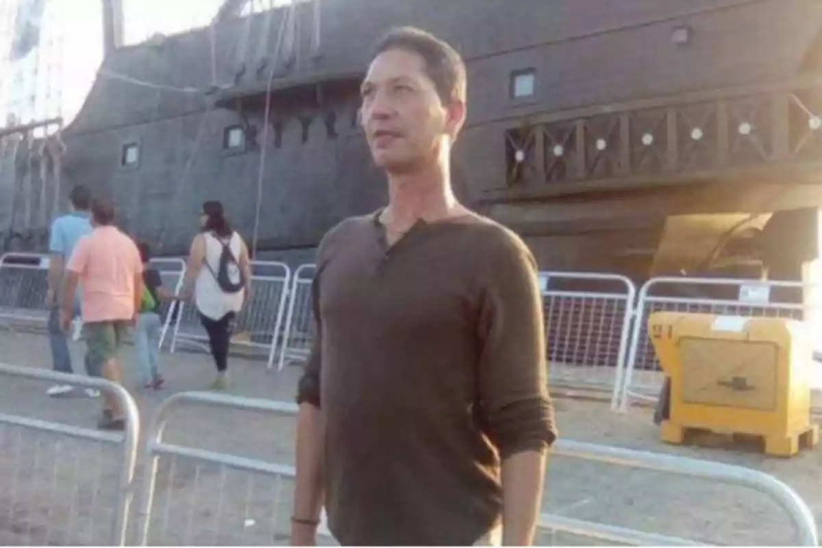 Un hombre de pie frente a una estructura que parece un barco antiguo, con varias personas caminando en el fondo.