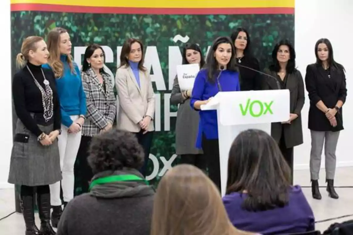 Un grupo de mujeres está de pie en un escenario, una de ellas está hablando en un podio con el logotipo de VOX, mientras las demás la escuchan atentamente.