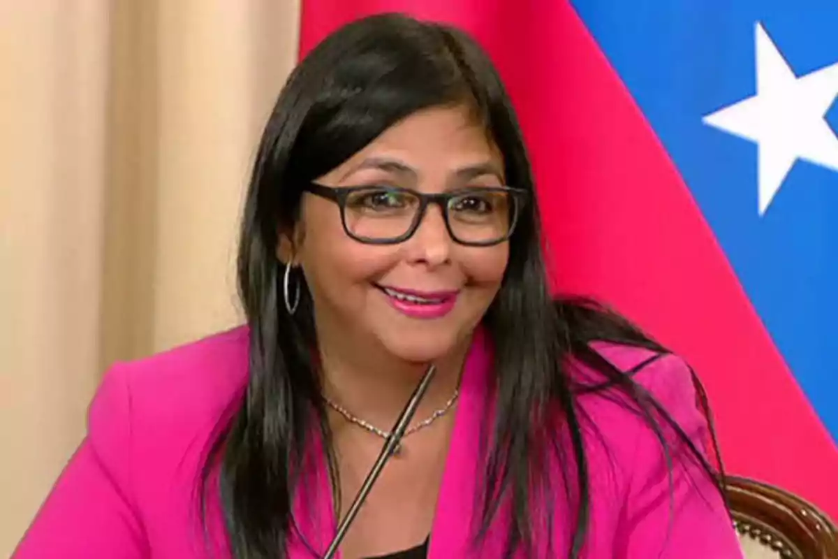 Mujer con gafas y chaqueta rosa sonriendo frente a un micrófono con una bandera de fondo.